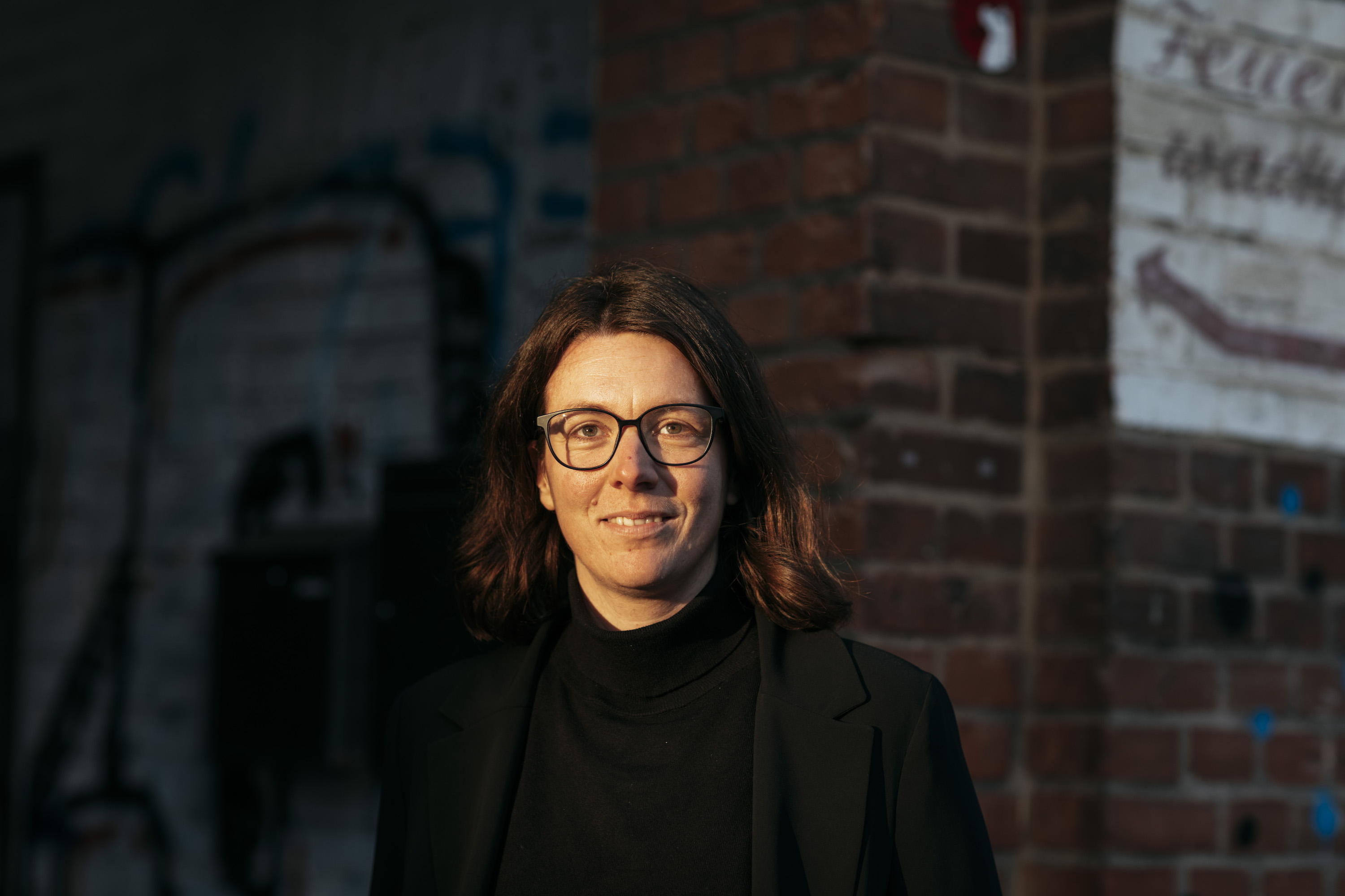 Portraitbild von Katrin Maiwald. Sie ist weiß gelesen, hat halblanges braunes Haar, trägt eine Brille und ein schwarzes Jackett. Sie richtet den Blick in die Ferne und lächelt. Im Hintergrund ist eine dunkelrote Backsteinwand zu sehen.